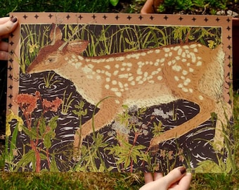 May include: A framed print featuring a fawn in a marshy area. The fawn has brown fur with white spots and is surrounded by green plants and flowers. The artwork has a brown border with star-like designs.