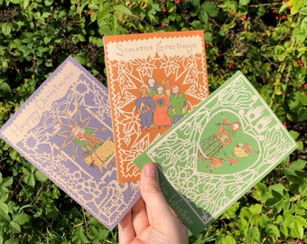 May include: Three Christmas cards with different designs. The first card is purple and features a person with a long stick and a star on top. The second card is orange and features a group of people in a star-shaped frame. The third card is green and features a person with a bird and a heart-shaped frame. The text on the cards reads "Happy Christmas", "Seasons Greetings", and "Warm Wishes".