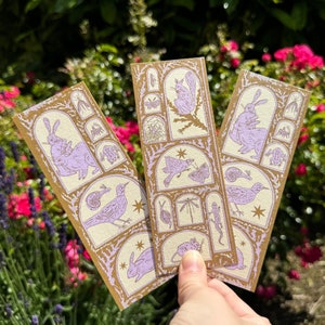 May include: Three rectangular bookmarks with a light brown background and a gold border. Each bookmark features a series of lavender illustrations of animals and insects. The bookmarks are held in front of a green and pink floral background.