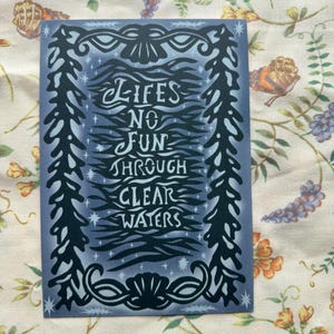 May include: A rectangular card with a blue background and white text that reads "LIFES NO FUN THROUGH CLEAR WATERS." The text is surrounded by a decorative black border with seashell and plant-like designs.