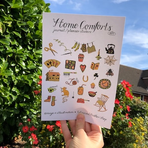 Puede incluir: Una hoja de pegatinas coloridas con ilustraciones de objetos hogare&ntilde;os como una tetera, una mecedora, una casa y una bola de lana. La hoja se titula "Home Comforts Journal/Planner Stickers 001" e incluye el nombre de usuario de las redes sociales @maya.c.illustration.