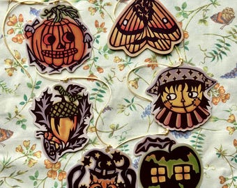 May include: A collection of Halloween-themed ornaments. The ornaments include a jack-o'-lantern, a moth, a witch, an acorn, a cat wearing a witch hat, and an apple with a house design. Each ornament is strung with a gold string.