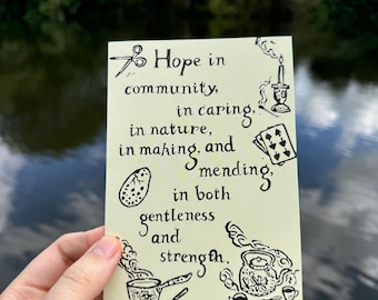May include: A yellow card with black ink illustrations and text that reads "Hope in community, in caring, in nature, in making, and mending, in both gentleness and strength."