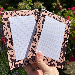 May include: Two pink notepad memo pads with a black butterfly and floral design border. The pads have a grid pattern.