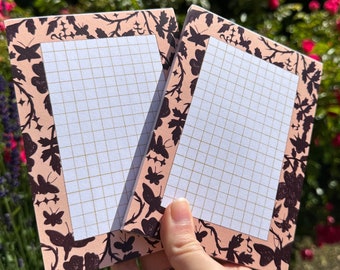 May include: Two pink notepad memo pads with a black butterfly and floral design border. The pads have a grid pattern.