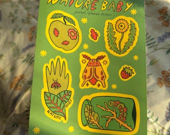 May include: A green sticker sheet with the words "NATURE BABY" and various nature-themed illustrations in yellow and orange. Includes images of a hand with a ladybug, a moth, and a dandelion. The text "journal/planner stickers" is also present.