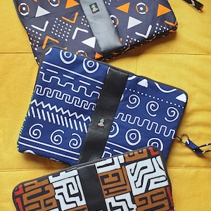 以下が含まれることがあります： 異なる幾何学模様の3つのラップトップスリーブ。スリーブは、黒い革のフラップとジッパー留め具が付いたファブリック製です。パターンには、オレンジ、ブルー、ブラウンの幾何学模様が含まれています。