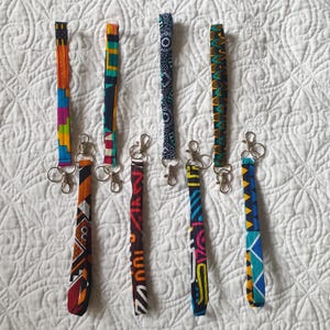 May include: Seven colorful patterned fabric keychains with silver key rings. The keychains are made of fabric with different geometric patterns in shades of blue, green, yellow, orange, red, black, and white.