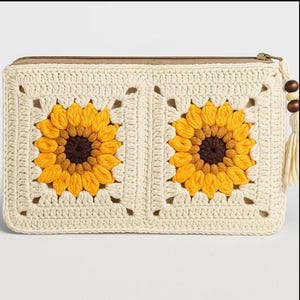 Könnte beinhalten: Cremefarbene Häkeltasche mit zwei Sonnenblumenmustern in Gelb und Braun. Die Tasche hat einen Reißverschluss und eine Quaste mit Holzperlen. Ein handgefertigtes Accessoire.