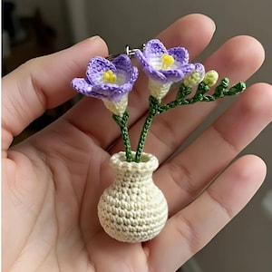 May include: A handmade crochet brooch featuring two purple and yellow flowers in a small cream-colored vase. The flowers have detailed petals and green stems. The vase is also crocheted, and the brooch has a pin on the back.