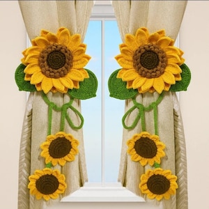 May include: Crocheted sunflower curtain tiebacks. The tiebacks feature large yellow sunflowers with brown centers and green leaves. Two smaller sunflowers hang below, connected by green yarn. The curtains are a neutral color, and the window is visible in the background.