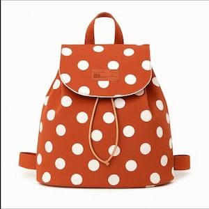 Könnte beinhalten: Rostfarbener Rucksack mit großen weißen Polka-Dots. Er hat einen Klappenverschluss, einen Kordelzug und einen oberen Griff. Die Tasche hat verstellbare Schultergurte und ein kleines braunes Etikett auf der Klappe.