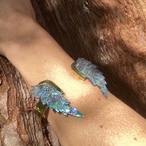 May include: Two gold bracelets with iridescent blue and green wings. The bracelets are worn on a person's arm.