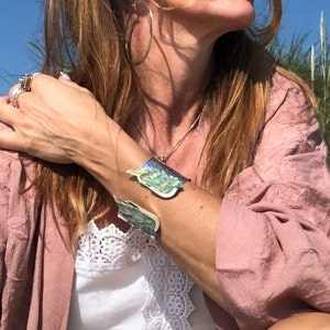 May include: A woman wearing a pink, embroidered top with a white lace trim and a silver bracelet with a green and blue iridescent design.