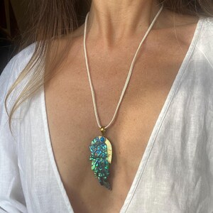 May include: A white cord necklace with a large iridescent green and blue abalone shell pendant shaped like a wing. The pendant has a gold accent and two small blue stones.