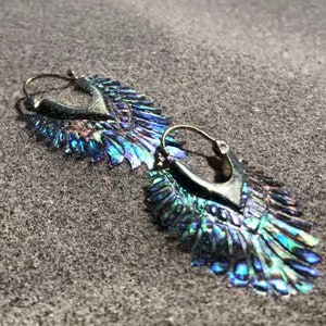 May include: A pair of hoop earrings featuring a V-shaped metal top and a dangling, fan-shaped fringe made of iridescent, multi-colored shell segments that shimmer with blue, green, and purple hues when catching the light.