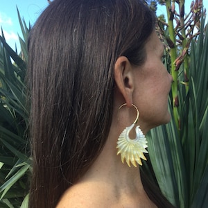 May include: A pair of large, gold hoop earrings with a white and yellow shell design. The earrings are worn by a person with long brown hair.