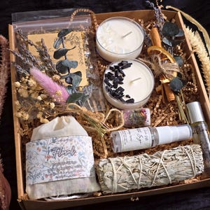 May include: A gift box containing two white candles, one topped with clear crystals and one with black stones, a floral sachet, a white sage smudge stick, a spray bottle labelled White Sage, a small glass vial of herbs, and dried botanical stems.