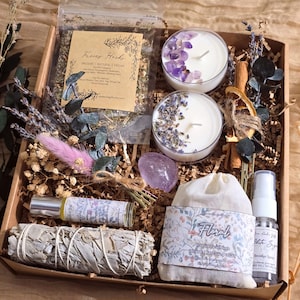 May include: A gift box containing two candles topped with amethyst and lavender, a bag of dried herbs labelled 'Fierce Herbs', a sage smudge stick, a floral-labelled pouch, a spray bottle, a rollerball, a polished amethyst stone, and dried flowers.