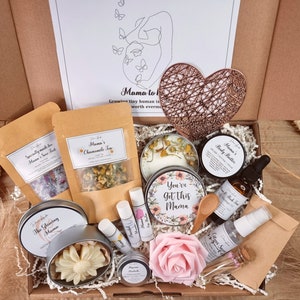 May include: A gift box containing two kraft paper pouches of tea and bath salts, a copper wire heart, a candle with floral labels reading You've Got This Mama, body butter, massage oil, a spray bottle, lip balms, a wooden spoon, and a pink rose.