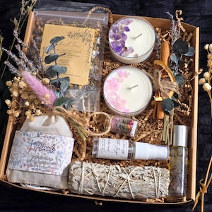 May include: A gift box containing two candles topped with amethyst and rose quartz crystals, a bag of loose leaf tea, a small glass vial of dried flowers, a spray bottle, a roller bottle, a sage smudge stick, and dried floral sprigs.