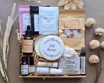 May include: A gift box containing various self-care items including three small pouches of clay, a bag of rose bath salts, a round tin labeled Just Because, a glass dropper bottle, a cuticle oil, a lip balm, a spray bottle, and a floral soap bar.