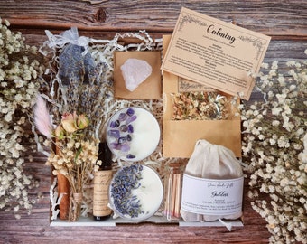 Puede incluir: Una caja de regalo blanca llena de artículos para la relajación y el cuidado personal. La caja incluye dos velas, una pequeña bolsa de hierbas secas, un cristal, una botella de aceite esencial, una pequeña bolsa de sales de baño y un ramo de flores secas. La caja está etiquetada como "Calming" y "Goddess".