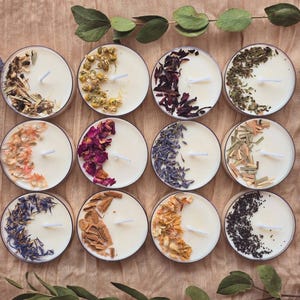 May include: Twelve round tea light candles, each with a white wax base and a different selection of dried herbs and flowers. The candles are displayed on a neutral surface, with lavender sprigs and greenery adding a natural aesthetic.
