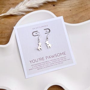 May include: Silver dog-shaped drop earrings with a heart cutout, presented on a white card that reads "YOU'RE PAWSOME." The card is set on a white tray with a decorative plant. A lovely accessory for dog lovers.