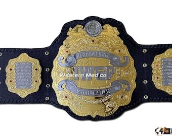 Pode incluir: Um cinturão de campeonato dourado e prateado com o texto "HEAVYWEIGHT CHAMPION" e "IWGP CHAMPION". O cinturão tem uma correia de couro preta e placas douradas decorativas com detalhes vermelhos. A imagem também inclui o texto "Western Med co".