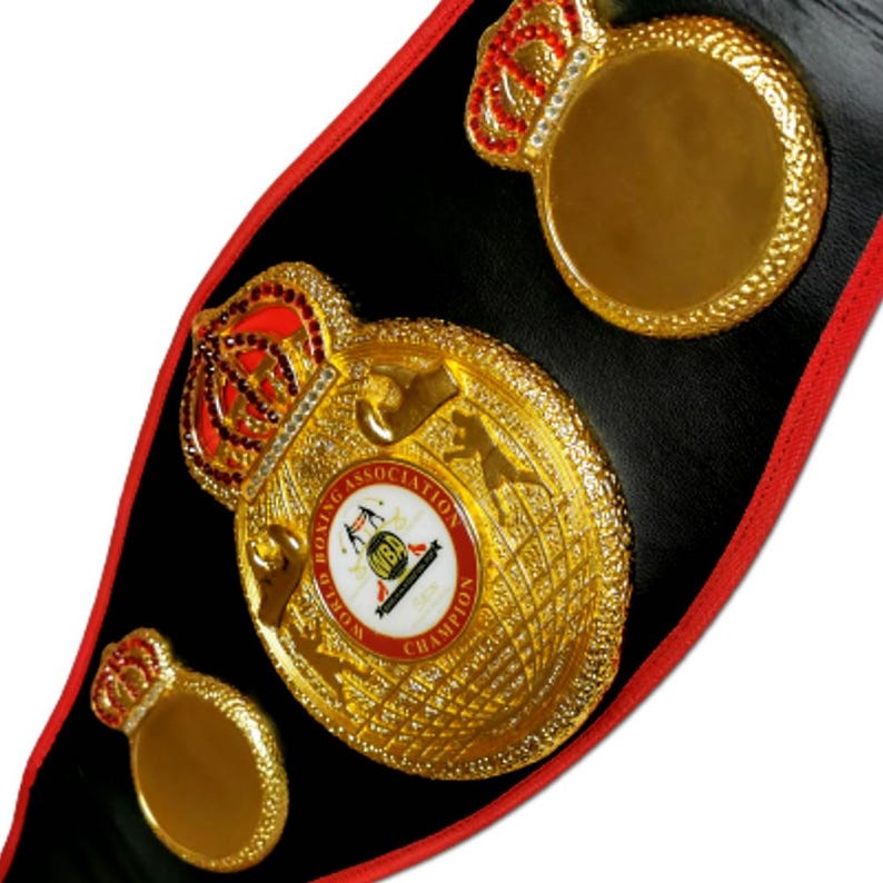 May include: A gold boxing championship belt with a black leather strap and red trim. The belt features a large central plate with a crown, the words "World Boxing Association" and "Champion", and a globe design. The belt also has two smaller plates with crowns.