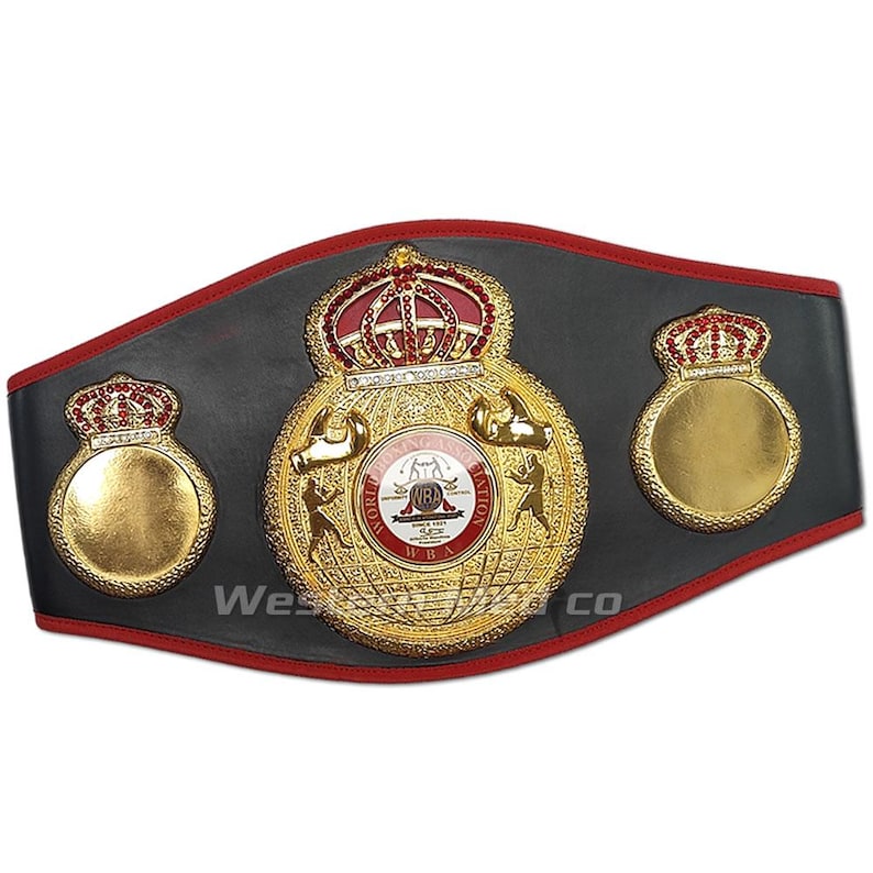 May include: A black and gold boxing championship belt with red accents. The belt features a large central emblem with the words "World Boxing Association" and "WBA" in gold lettering. The emblem also includes a crown, two boxing gloves, and a globe. The belt has two smaller crowns on either side of the central emblem.
