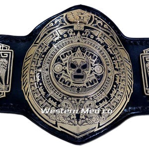 Puede incluir: Un cinturón de campeonato de lucha libre negro y dorado con intrincados diseños de inspiración azteca. La placa central presenta un diseño de máscara y las placas laterales muestran figuras de águilas. El cinturón está hecho de cuero negro.