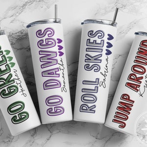 May include: Four white stainless steel tumblers with various phrases and names printed on them. The phrases include "GO GREEN", "GO DAWGS", "ROLL SKIES", and "JUMP AROUND". Each tumbler has a different color scheme and heart accents.
