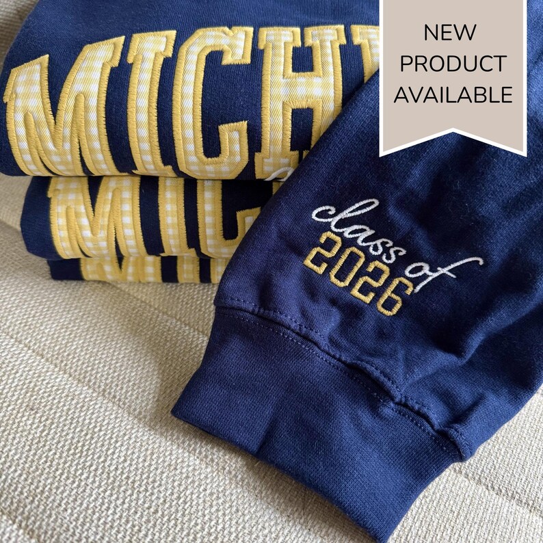 May include: Navy blue sweatshirts with yellow gingham-style lettering that reads "MICHIGAN". One sleeve features "class of 2026" in embroidered script. A "NEW PRODUCT AVAILABLE" tag is visible.