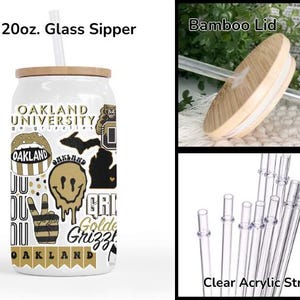 K&ouml;nnte beinhalten: Ein wei&szlig;er 591 ml Glasbecher mit Bambusdeckel und klarem Acrylstrohhalm. Der Becher hat Aufkleber mit "OAKLAND UNIVERSITY" und "Golden Grizz". Das Design umfasst einen B&auml;ren, ein Smiley und ein Friedenszeichen.