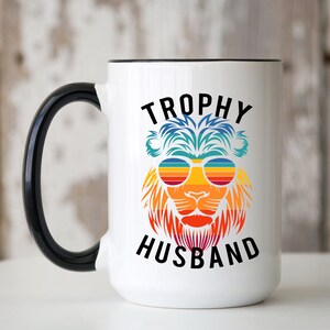 May include: White ceramic coffee mug with a black handle and rim. The mug features a colorful graphic of a lion wearing sunglasses with the text "TROPHY HUSBAND" above and below the lion.