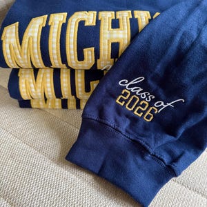 May include: Navy blue sweatshirts with the word "MICHIGAN" in yellow gingham letters. The sleeve of one sweatshirt displays "class of 2026" in white and yellow embroidery. The sweatshirts are folded on a neutral-colored surface.
