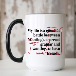 May include: White coffee mug with a black handle. The mug has a red pencil and red markings on it. The text on the mug reads: "My life is a constant battle between wanting to correct grammar and wanting to have friends."