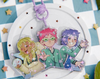 Może przedstawiać: Przezroczysty brelok akrylowy z trzema postaciami anime o różowych, zielonych i fioletowych włosach. Postacie stoją na tle w kształcie gwiazdy z efektem błyszczenia. Brelok ma fioletowy metalowy pierścień.
