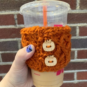 Puede incluir: Un calentador de taza de café de punto de ganchillo marrón con dos botones de madera con forma de calabaza. El calentador está en una taza de plástico transparente con una pajita.