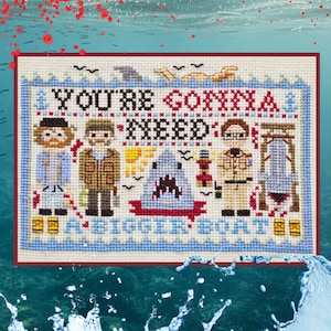 Puede incluir: Un bordado de punto de cruz sobre tela blanca con tres figuras, un tiburón, un faro y un barco. El texto dice "YOU'RE GONNA NEED A BIGGER BOAT" en hilo negro y azul. El diseño incluye detalles en rojo y un borde de color azul.