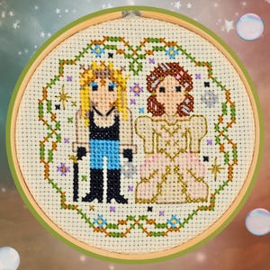 May include: A cross-stitch embroidery hoop featuring two cartoon figures, one with blonde hair and the other with brown hair, surrounded by a green and gold floral border. The figures are dressed in fantasy-inspired clothing, with a black top and blue pants for the first figure and a pink dress for the second.