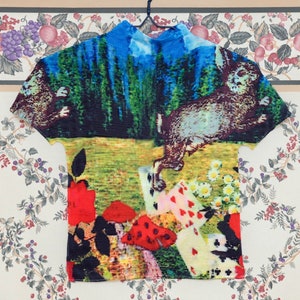Puede incluir: Una camisa de manga corta con un estampado colorido que presenta un conejo, cartas de juego y flores. La camisa tiene un cuello alto y está hecha de una tela plisada.