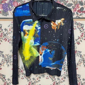 May include: Black long-sleeve button-down shirt with a unique, colorful print. The design features abstract art with yellow, blue, and orange hues, and a celestial theme. The shirt has a pleated texture.