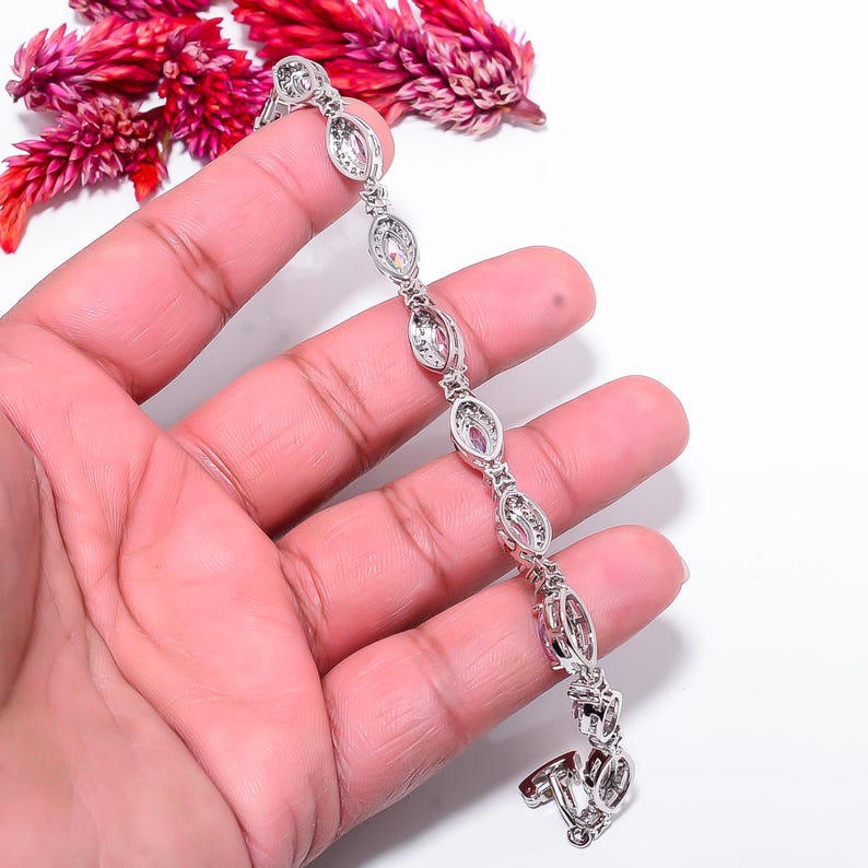 Pode incluir: Uma pulseira de prata com pedras preciosas ovais e transparentes. A pulseira &eacute; segurada em uma m&atilde;o, com fundo branco e flores vermelhas no canto superior esquerdo. Fechamento por fecho.