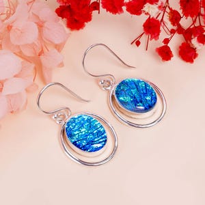 Könnte beinhalten: Ein Paar silberne Ohrringe mit blauen, schillernden, ovalen Glassteinen, die in einem Doppelkreisdesign gefasst sind.
