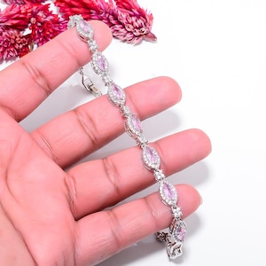 Pode incluir: Uma pulseira de prata com pedras rosas ovais, cada uma rodeada por pedras claras menores. A pulseira &eacute; exibida sobre um fundo claro, com flores rosas no canto superior esquerdo.