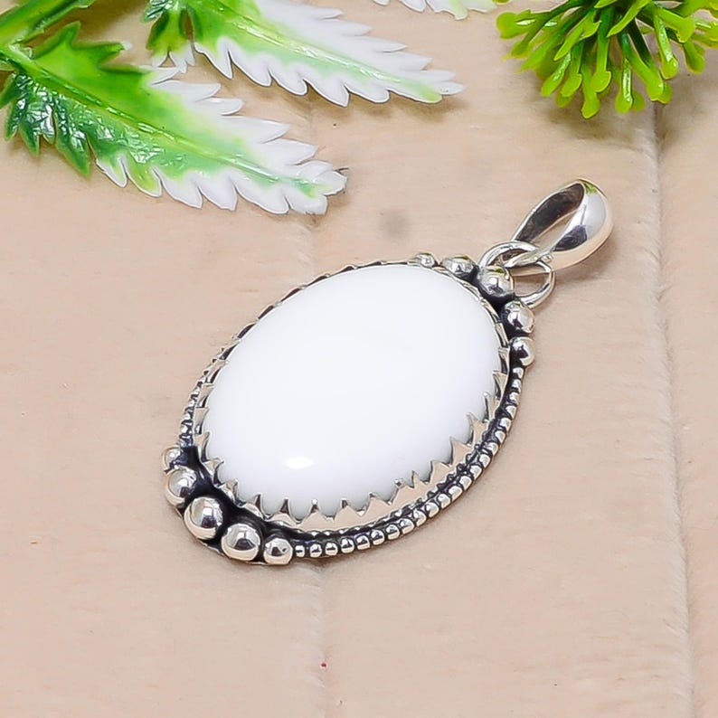 Puede incluir: Colgante ovalado blanco con una fianza plateada y bordes decorativos. El colgante tiene un centro de piedra blanca lisa y pulida, rodeado por un borde plateado detallado con peque&ntilde;os acentos redondos. El dise&ntilde;o es simple pero elegante.