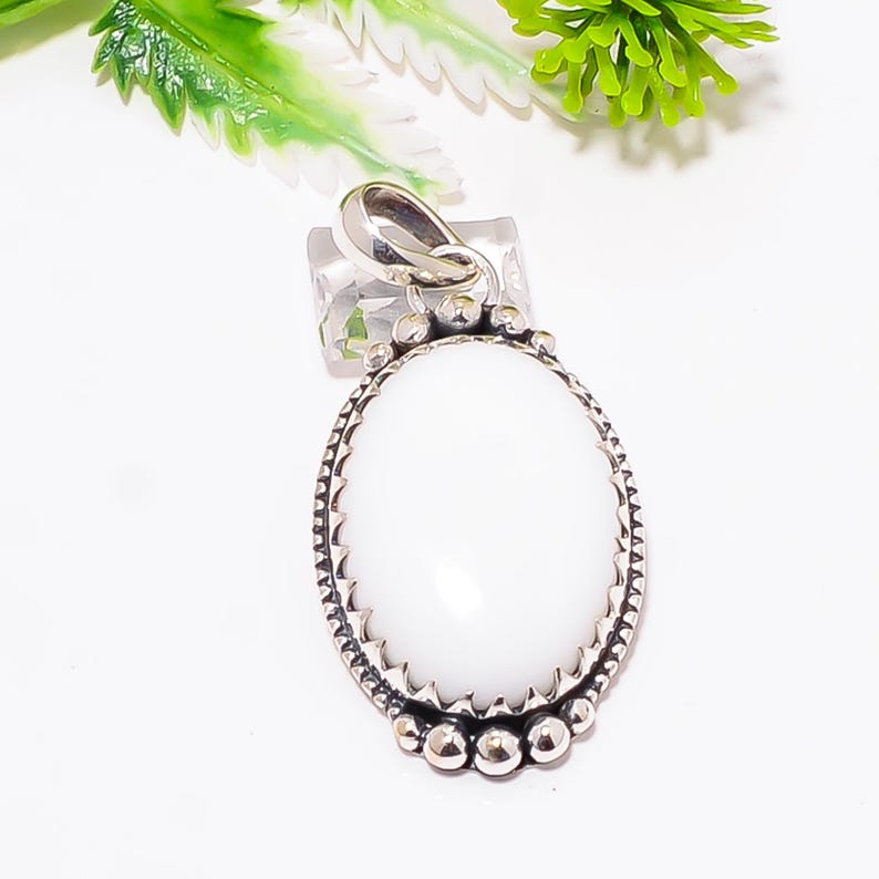 Puede incluir: Colgante ovalado de piedra preciosa blanca con un engaste de metal plateado. El colgante tiene un borde texturizado y un lazo para una cadena. La piedra preciosa es lisa y pulida. Es una joya.